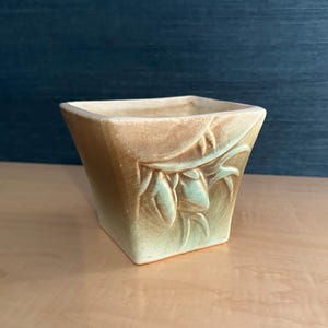 Puede incluir: Una maceta cuadrada de cerámica con un degradado marrón claro y verde. La maceta presenta un diseño floral en relieve en los laterales. El interior es de un tono marrón más claro. La maceta está sobre una superficie marrón claro.