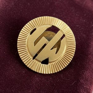 May include: A gold-colored, circular pin with a ridged outer edge and the letters "GW" in the center. The pin is set against a dark burgundy velvet background.
