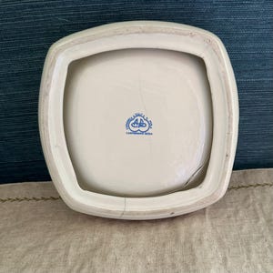 Pode incluir: Um prato quadrado de cer&acirc;mica branca com uma borda ligeiramente curva. A parte inferior do prato tem um emblema azul e o texto "CERAMICA ITALY P.A. POLA CARPIGNANO SESIA". O prato tem uma rachadura.