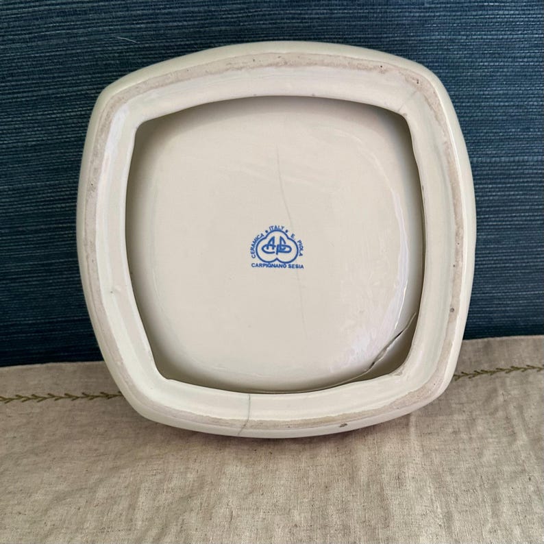 Pode incluir: Um prato quadrado de cer&acirc;mica branca com uma borda ligeiramente curva. A parte inferior do prato tem um emblema azul e o texto "CERAMICA ITALY P.A. POLA CARPIGNANO SESIA". O prato tem uma rachadura.