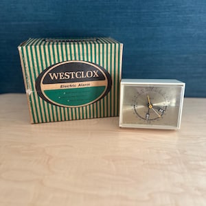 以下が含まれることがあります： ヴィンテージのウェストクロックス電気目覚まし時計とオリジナルの箱。時計は白で、金色の文字盤に黒い数字と針が付いています。箱は緑色の縦縞模様で、ウェストクロックスのロゴが入っています。時計の幅は約10cmです。
