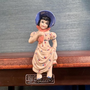 Puede incluir: Figurita de porcelana vintage de una mujer con un sombrero azul, una bufanda melocotón y un vestido floral. La figura está sobre una superficie marrón con un letrero metálico "Hi-L by ALS".