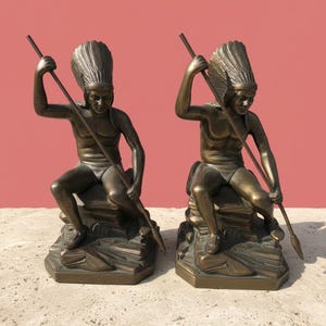 May include: Two bronze statues depict Native American figures seated on rock formations, each holding a spear. The figures wear headdresses and are posed with their spears raised. The statues have a detailed, antique finish and are set against a pink background.