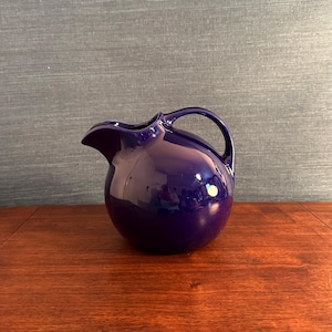 May include: A deep cobalt blue ceramic pitcher with a rounded, spherical body, a wide pouring spout, and a curved handle. The surface of the pitcher is smooth and glossy, reflecting light across its curved form, creating a bright highlight.