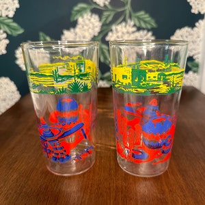 May include: Two clear glass tumblers with a Southwestern desert scene printed on them. The scene features a yellow sky, green cacti, red adobe buildings, and blue figures.