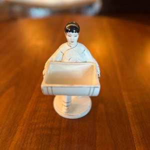 May include: A white porcelain figurine of a person with black hair, wearing a white garment with thin gold trim. The figure holds a square, open-topped white container with gold-lined edges in front of them, supported by a circular base.