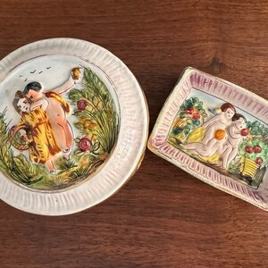 Puede incluir: Dos platos de cer&aacute;mica con escenas pintadas de personas en un entorno de jard&iacute;n. Un plato es redondo y presenta una pareja en un jard&iacute;n con &aacute;rboles y flores. El otro plato es rectangular y presenta a tres personas en un jard&iacute;n con &aacute;rboles frutales y flores.