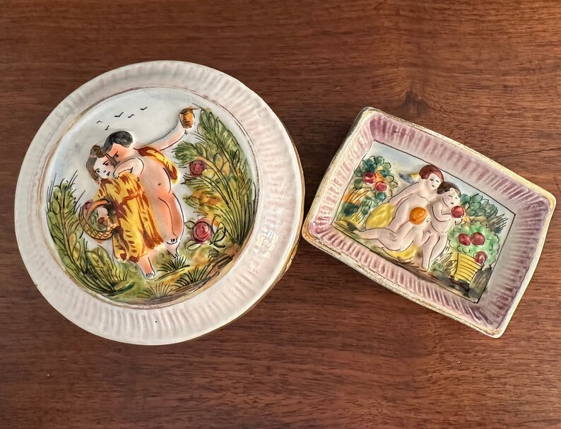 Puede incluir: Dos platos de cer&aacute;mica con escenas pintadas de personas en un entorno de jard&iacute;n. Un plato es redondo y presenta una pareja en un jard&iacute;n con &aacute;rboles y flores. El otro plato es rectangular y presenta a tres personas en un jard&iacute;n con &aacute;rboles frutales y flores.
