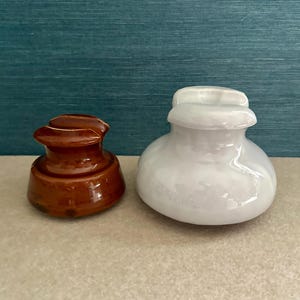 May include: Two ceramic insulators, one brown and one white, are displayed. The brown insulator has a glossy finish. The white insulator has a rounded body and a flat top. Both are on a neutral-toned surface against a teal background.