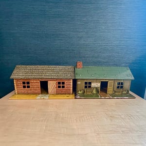 Può includere: Due edifici giocattolo vintage affiancati. A sinistra, una capanna di tronchi marrone con un tetto grigio testurizzato. A destra, un edificio verde e marrone chiaro con l'etichetta Headquarters U.S. Army Training Center, camino e finestre.