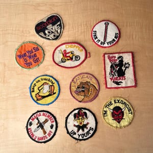 May include: A collection of embroidered patches with various designs and text. The patches feature phrases like "Love You," "Chopper," and "Keep on Truckin'." Other patches depict a bulldog, a devil, and a jester. The patches are in various shapes and colors.