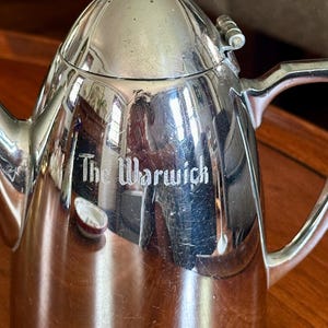 May include: A polished silver-tone metal coffee pot with a hinged lid, a curved handle, and a spout. The body of the pot features the engraved text The Warwick. The surface is reflective and shows light surface scratches throughout the metal finish.