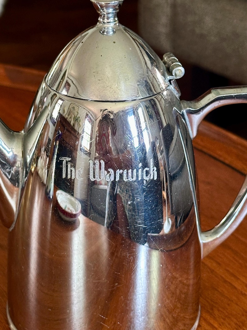 May include: A polished silver-tone metal coffee pot with a hinged lid, a curved handle, and a spout. The body of the pot features the engraved text The Warwick. The surface is reflective and shows light surface scratches throughout the metal finish.