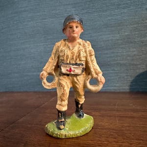 May include: A painted cast iron figurine of a soldier wearing a helmet and camouflage uniform. The soldier is holding two rings in his hands and has a red cross on his belt.
