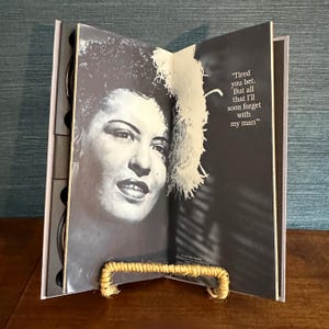 May include: A black and white photograph of a woman with text that reads "Tired you bet. But all that I'll soon forget with my man." The photograph is displayed in a book-like format, propped up by a woven stand.