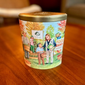 Puede incluir: Una lata metálica cilíndrica con tapa dorada que presenta una ilustración colorida de personas cosechando productos en un jardín. El diseño incluye el logotipo de Girl Scouts, un letrero que dice Food Bank Harvest y varias figuras con cestas de verduras.