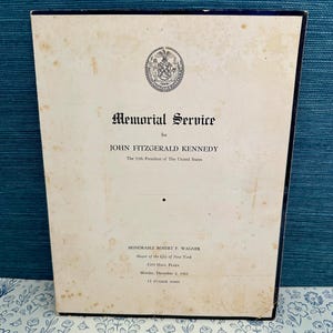 May include: A vintage memorial service program for John F. Kennedy, the 35th President of the United States. The program is cream-colored with black text and a New York City seal. The service was held on December 2, 1963.