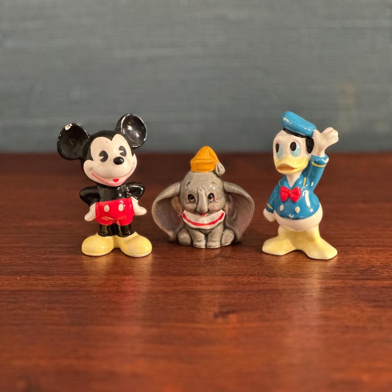 K&ouml;nnte beinhalten: Drei Disney-Keramikfiguren: Micky Maus in Schwarz, Rot und Gelb; Dumbo der Elefant in Grau und Gelb; und Donald Duck in Blau, Wei&szlig; und Gelb. Die Figuren sind auf einer dunkelbraunen Oberfl&auml;che ausgestellt.
