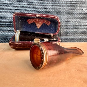 May include: An antique brown glass pipe with a metal rim and a separate black mouthpiece in a red velvet case. The case has a blue interior and a decorative emblem.