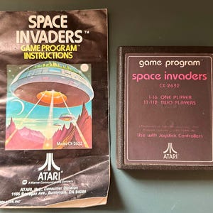 May include: A black Atari Space Invaders game cartridge and its matching instruction manual. The manual features a colorful illustration of a flying saucer over a landscape. The cartridge label displays the game title, model number CX-2632, and player options.