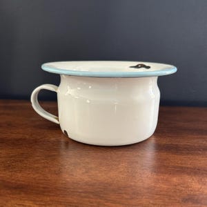 Op de afbeelding: Een witte emaille po met een lichtblauwe rand en handvat. De vintage pot heeft een klein handvat en een licht uitlopende rand. De pot staat op een houten oppervlak.
