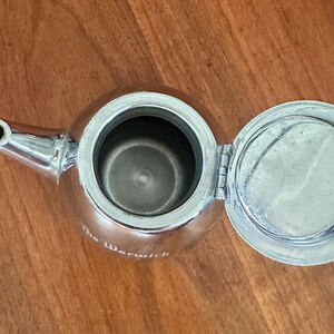 May include: A polished silver-tone metal teapot viewed from above with the lid open. The teapot features a curved spout and a circular opening. The text The Warwick is printed in white on the side of the metallic body of the teapot.
