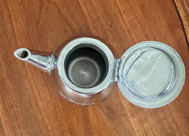 May include: A polished silver-tone metal teapot viewed from above with the lid open. The teapot features a curved spout and a circular opening. The text The Warwick is printed in white on the side of the metallic body of the teapot.