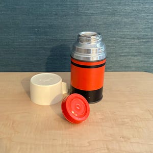 May include: A vintage thermos with a red and black body, silver top, and a white cup. The red lid is detached, and the thermos is on a light wood surface.