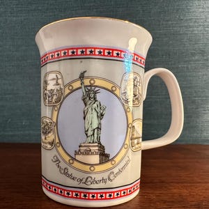Puede incluir: Taza de cerámica blanca con borde dorado y un diseño colorido con la Estatua de la Libertad y el texto "The Statue of Liberty Centennial".