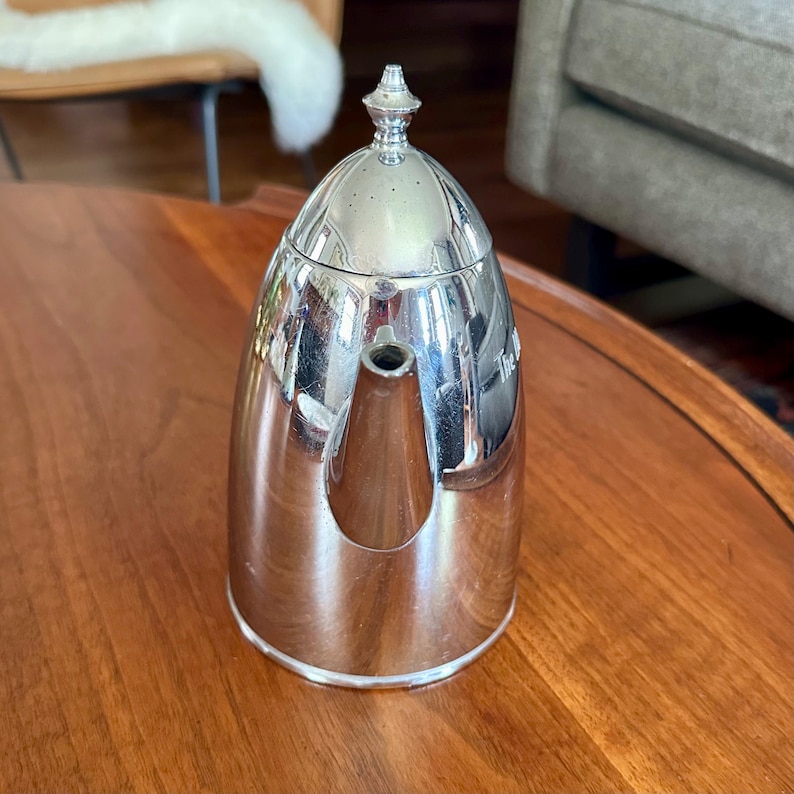May include: A polished silver-tone metal teapot with a tall, dome-shaped lid topped by a small finial. The teapot features a smooth, reflective surface, a tapered body, and a short, integrated spout extending from the front of the vessel.