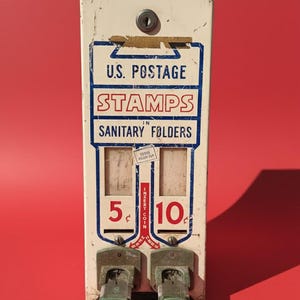 Puede incluir: Dispensador de sellos postales estadounidenses vintage con un exterior de metal pintado de blanco y azul. El dispensador tiene dos ranuras para sellos con un precio de 5¢ y 10¢. Se muestra el texto "U.S. Postage Stamps in Sanitary Folders".