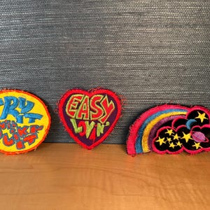 May include: Three colorful embroidered patches with text and designs. One is a yellow circle with blue text that reads "TRY IT YOU'LL LIKE IT". Another is a heart-shaped patch with "EASY LIVIN" in green and red. The third is a rainbow, clouds, and stars.