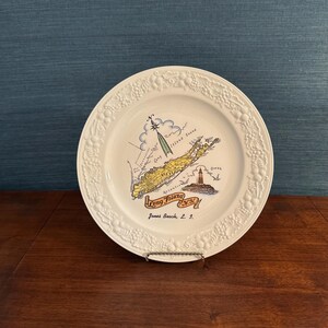 May include: A white ceramic decorative plate featuring a colorful map illustration of Long Island, N.Y. with a lighthouse, a compass rose, and the text Long Island N.Y. and Jones Beach, L.I. The plate has a raised embossed fruit and vine border.