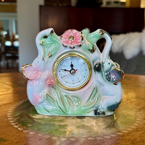 May include: A ceramic quartz clock with a floral and elephant design. The clock face is white with gold trim and black numbers. The clock is approximately 6 inches tall and features pink, green, and gold accents.