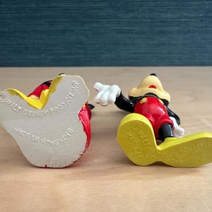 May include: Two vintage Mickey Mouse figurines. One is lying on its back, revealing the text "Walt Disney Productions Made in Hong Kong". The other is lying on its side, showing the bottom of its yellow shoe.