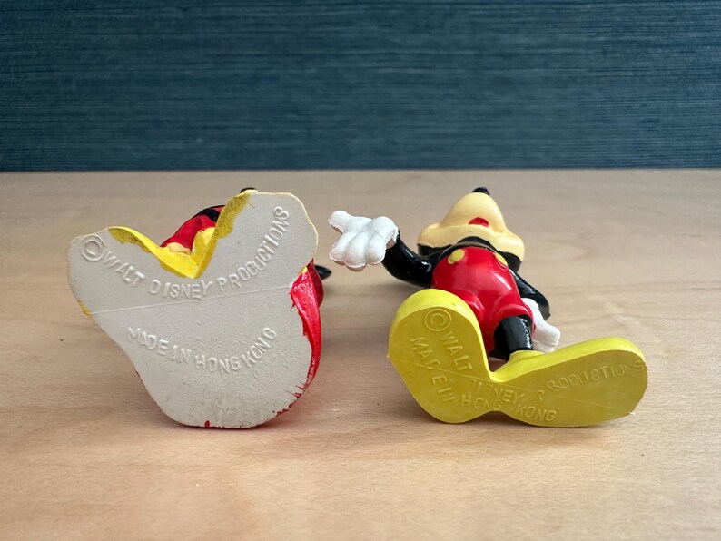 May include: Two vintage Mickey Mouse figurines. One is lying on its back, revealing the text "Walt Disney Productions Made in Hong Kong". The other is lying on its side, showing the bottom of its yellow shoe.