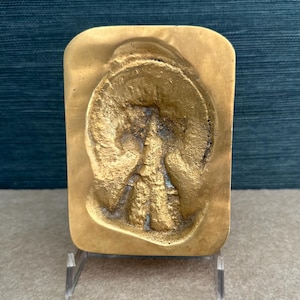 May include: A rectangular, gold-colored relief sculpture with a detailed, textured design. The relief features a central figure within a circular frame, possibly a portrait or emblem. The piece is displayed on a clear acrylic stand.