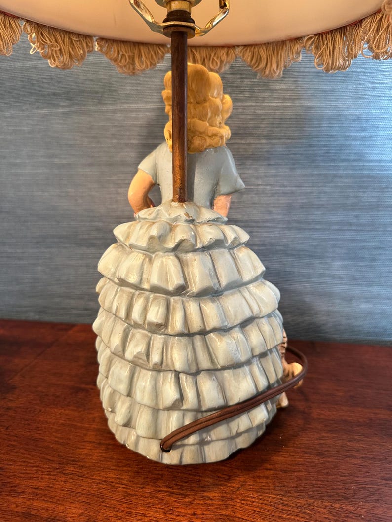 Puede incluir: Una l&aacute;mpara de mesa vintage con una figura de cer&aacute;mica de una mujer con un vestido azul y una falda con volantes. La l&aacute;mpara tiene una pantalla blanca con un borde de flecos.