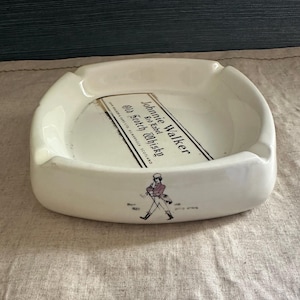 Pode incluir: Cinzeiro de cer&acirc;mica branca com design retangular. O cinzeiro apresenta o texto "Johnnie Walker Red Label Old Scotch Whisky" e um gr&aacute;fico de um homem a andar. O cinzeiro tem uma forma curva com uma pequena aba.