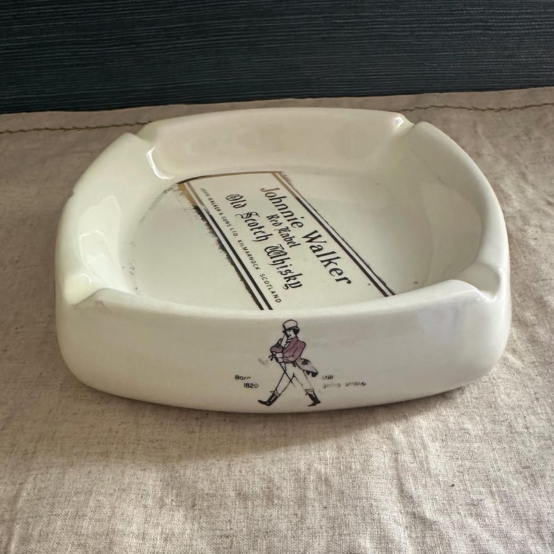 Pode incluir: Cinzeiro de cer&acirc;mica branca com design retangular. O cinzeiro apresenta o texto "Johnnie Walker Red Label Old Scotch Whisky" e um gr&aacute;fico de um homem a andar. O cinzeiro tem uma forma curva com uma pequena aba.
