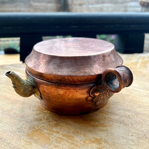 May include: A small, copper teapot with a lid and a spout shaped like a dragon's head. The teapot has a brass handle and decorative details.