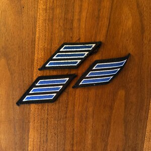 May include: Three parallelogram-shaped embroidered patches featuring a black border and three horizontal blue stripes separated by white lines. These fabric emblems are designed with a slanted orientation and clean, stitched edges for application.