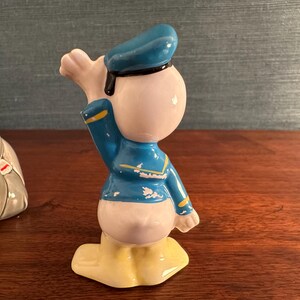 K&ouml;nnte beinhalten: Eine Keramikfigur von Donald Duck in Matrosenkleidung. Die Figur ist haupts&auml;chlich wei&szlig; mit einer blauen Jacke und M&uuml;tze, gelben F&uuml;&szlig;en und einer schwarzen Fliege. Auf einer Holzoberfl&auml;che.
