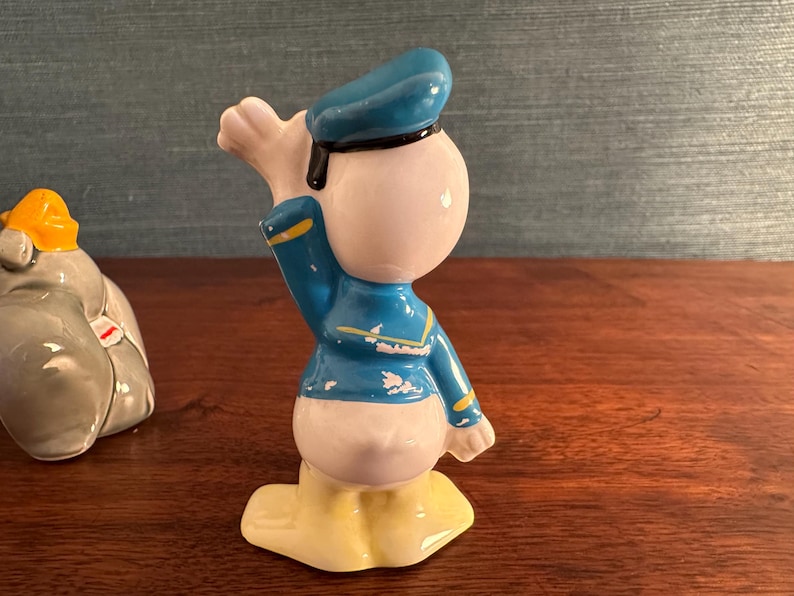 K&ouml;nnte beinhalten: Eine Keramikfigur von Donald Duck in Matrosenkleidung. Die Figur ist haupts&auml;chlich wei&szlig; mit einer blauen Jacke und M&uuml;tze, gelben F&uuml;&szlig;en und einer schwarzen Fliege. Auf einer Holzoberfl&auml;che.