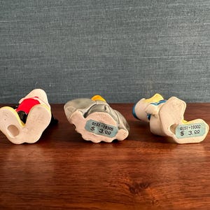 K&ouml;nnte beinhalten: Drei Keramikfiguren von Zeichentrickfiguren, jede in einer anderen Pose. Eine Figur tr&auml;gt ein rot-wei&szlig;es Outfit, eine andere ist grau mit einem gelben Hut, und die dritte tr&auml;gt einen blauen Hut und ein blaues Hemd. Die Figuren stehen auf einer Holzoberfl&auml;che.