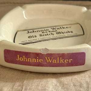 Pode incluir: Cinzeiro de cer&acirc;mica creme com formato retangular. O cinzeiro apresenta o texto "Johnnie Walker" em letras douradas em uma faixa roxa. O centro tem um r&oacute;tulo com texto sobre "Old Scotch Whisky".