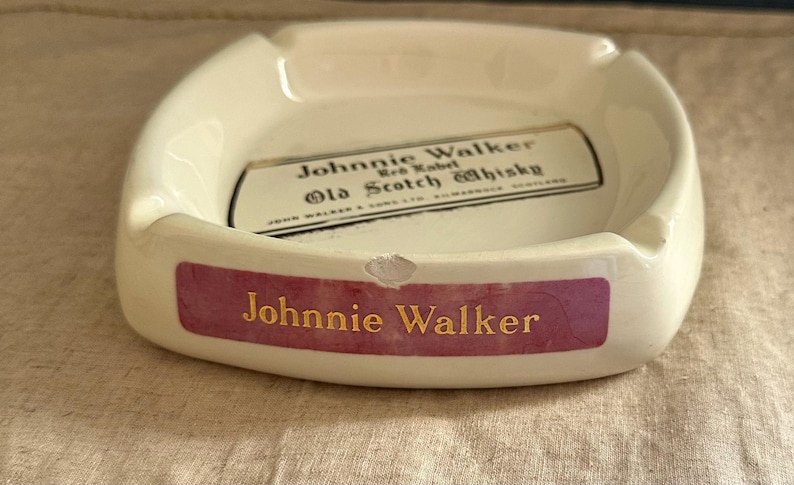 Pode incluir: Cinzeiro de cer&acirc;mica creme com formato retangular. O cinzeiro apresenta o texto "Johnnie Walker" em letras douradas em uma faixa roxa. O centro tem um r&oacute;tulo com texto sobre "Old Scotch Whisky".