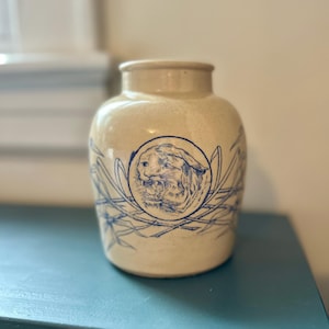 Puede incluir: Una vasija de cerámica color crema con una ilustración en tinta azul de una cabeza de tigre gruñendo dentro de un borde circular. El diseño incluye patrones de follaje tipo bambú que rodean el exterior del recipiente.