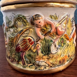 Puede incluir: Un tarro de cer&aacute;mica con tapa, decorado con una escena colorida de un querub&iacute;n, un p&aacute;jaro y flores. El tarro tiene un borde dorado alrededor de la parte superior e inferior.