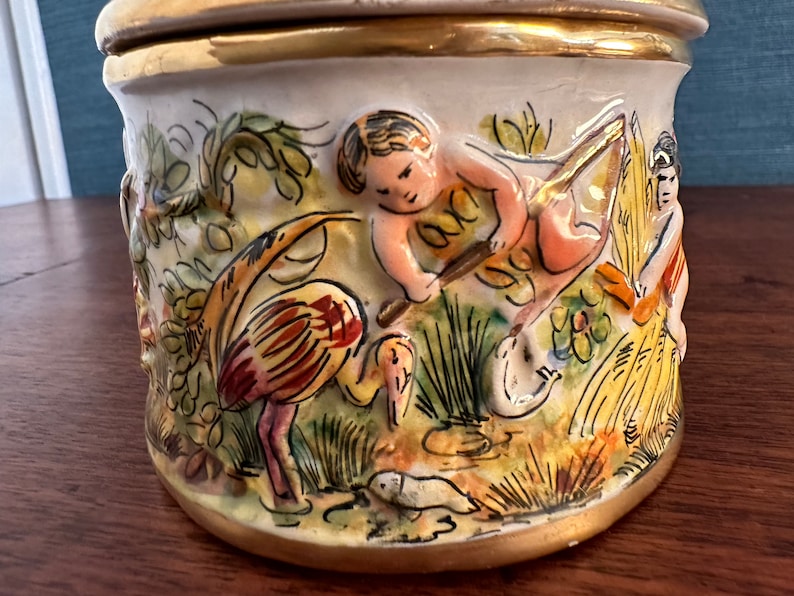 Puede incluir: Un tarro de cer&aacute;mica con tapa, decorado con una escena colorida de un querub&iacute;n, un p&aacute;jaro y flores. El tarro tiene un borde dorado alrededor de la parte superior e inferior.